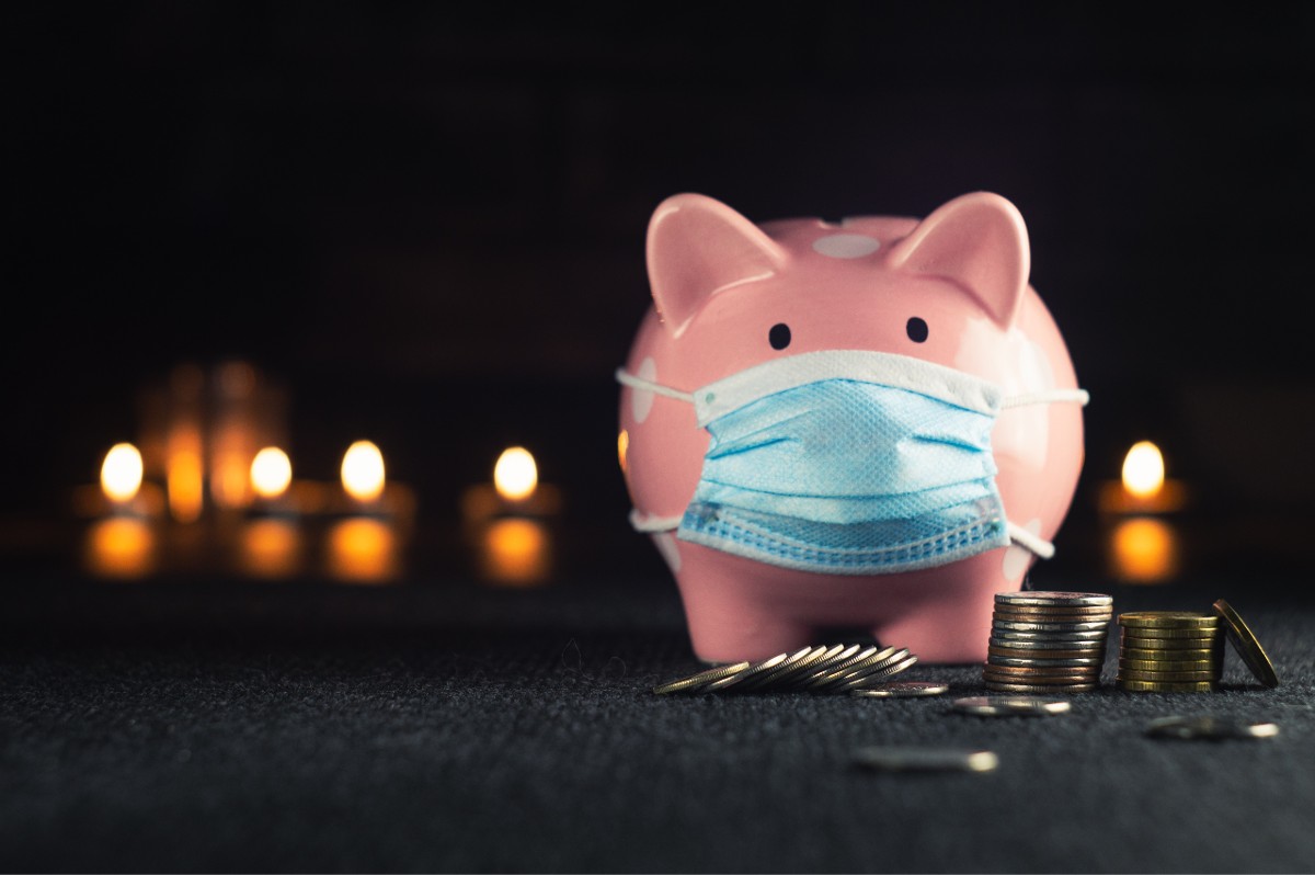 To illustrate the idea of health system profits, a pink piggy bank wearing a blue surgical mask is surrounded by piles of coins. Photo by Konstantin Evdokimov via Unsplash.