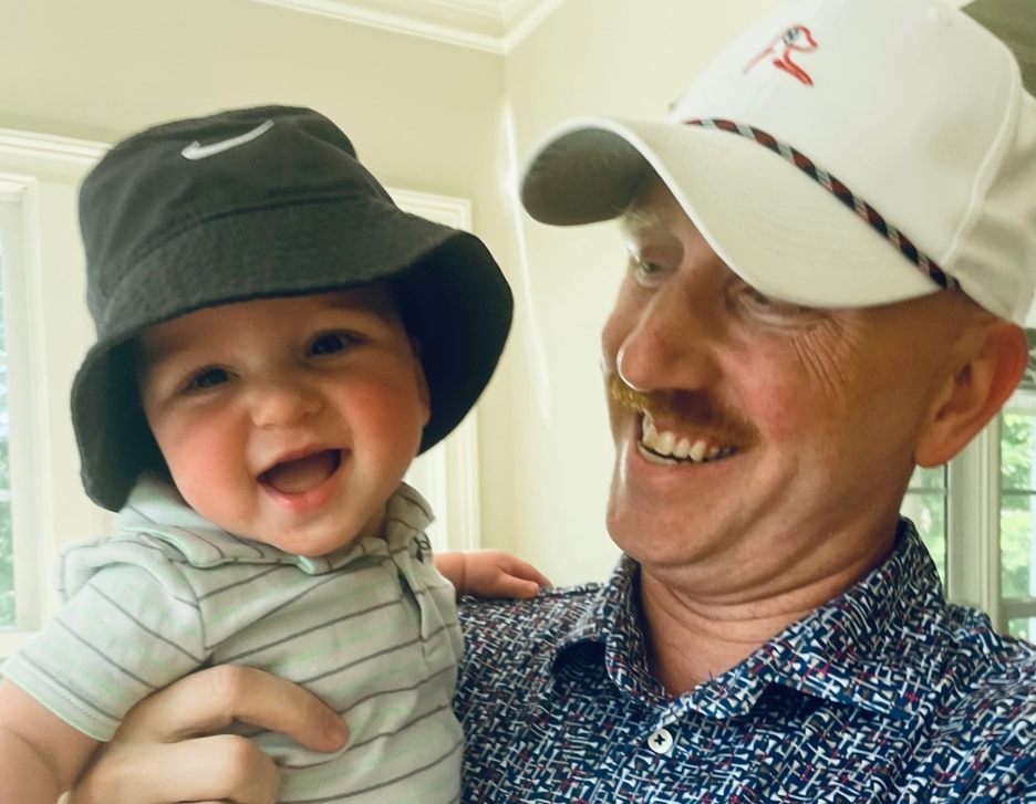 Greg Flicek, wearing a white golf hat, smiles with his infant son in a blue bucket hat