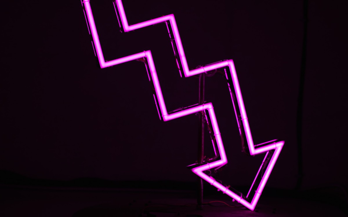 A down arrow in pink neon against a black background illustrates the idea of DRG downgrades. Photo by Ussama Azam on Unsplash.